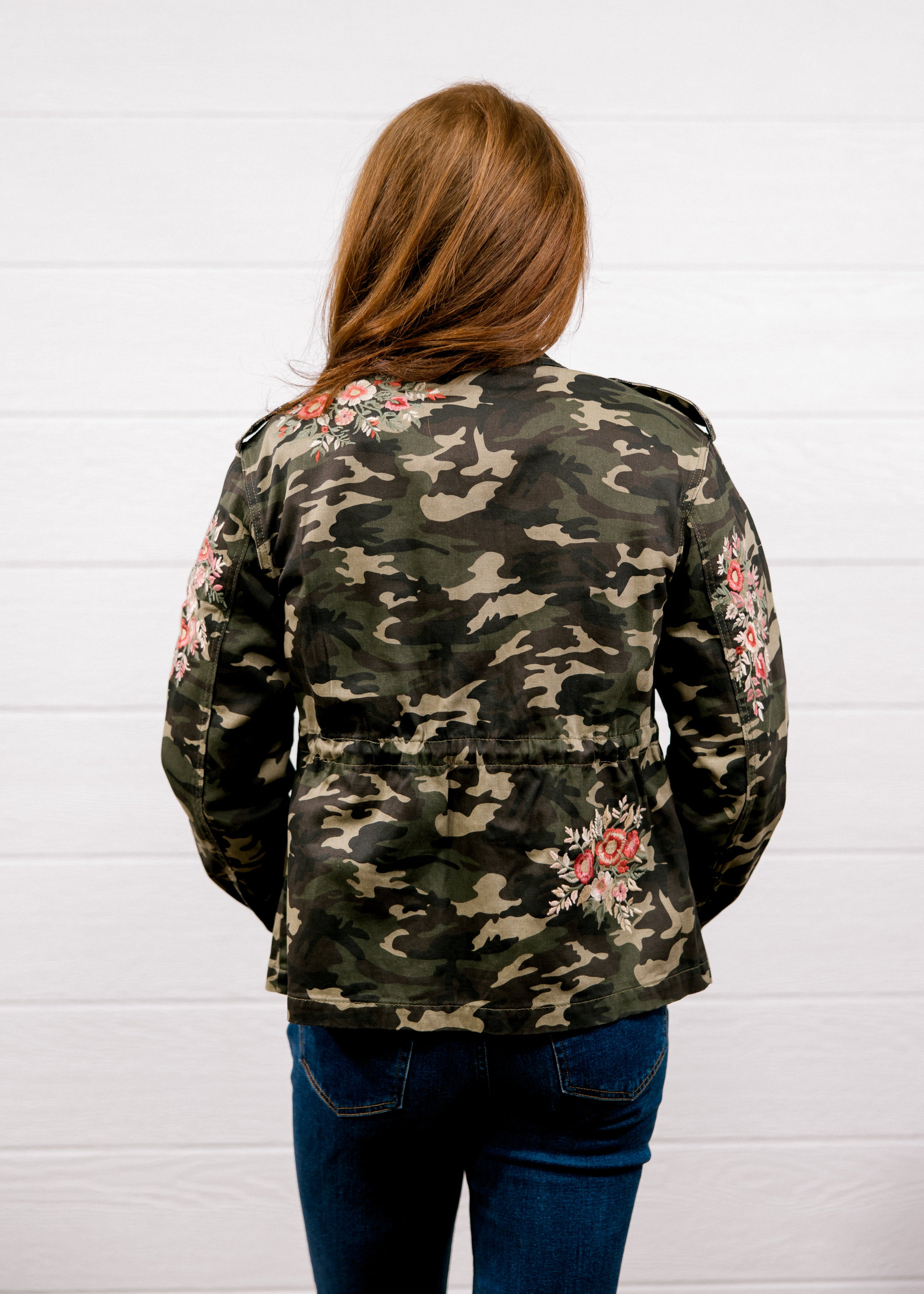 A woman viewed from behind wearing a camouflage military-style jacket with pink floral embroidery on the sleeves and back, paired with blue jeans, standing against a white wood-paneled wall.