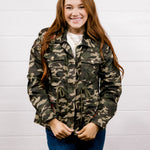 A smiling young woman with long red hair stands against a white paneled wall wearing a green and brown camouflage jacket over a white shirt and blue jeans.