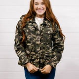 A smiling young woman with long red hair stands against a white paneled wall wearing a green and brown camouflage jacket over a white shirt and blue jeans.