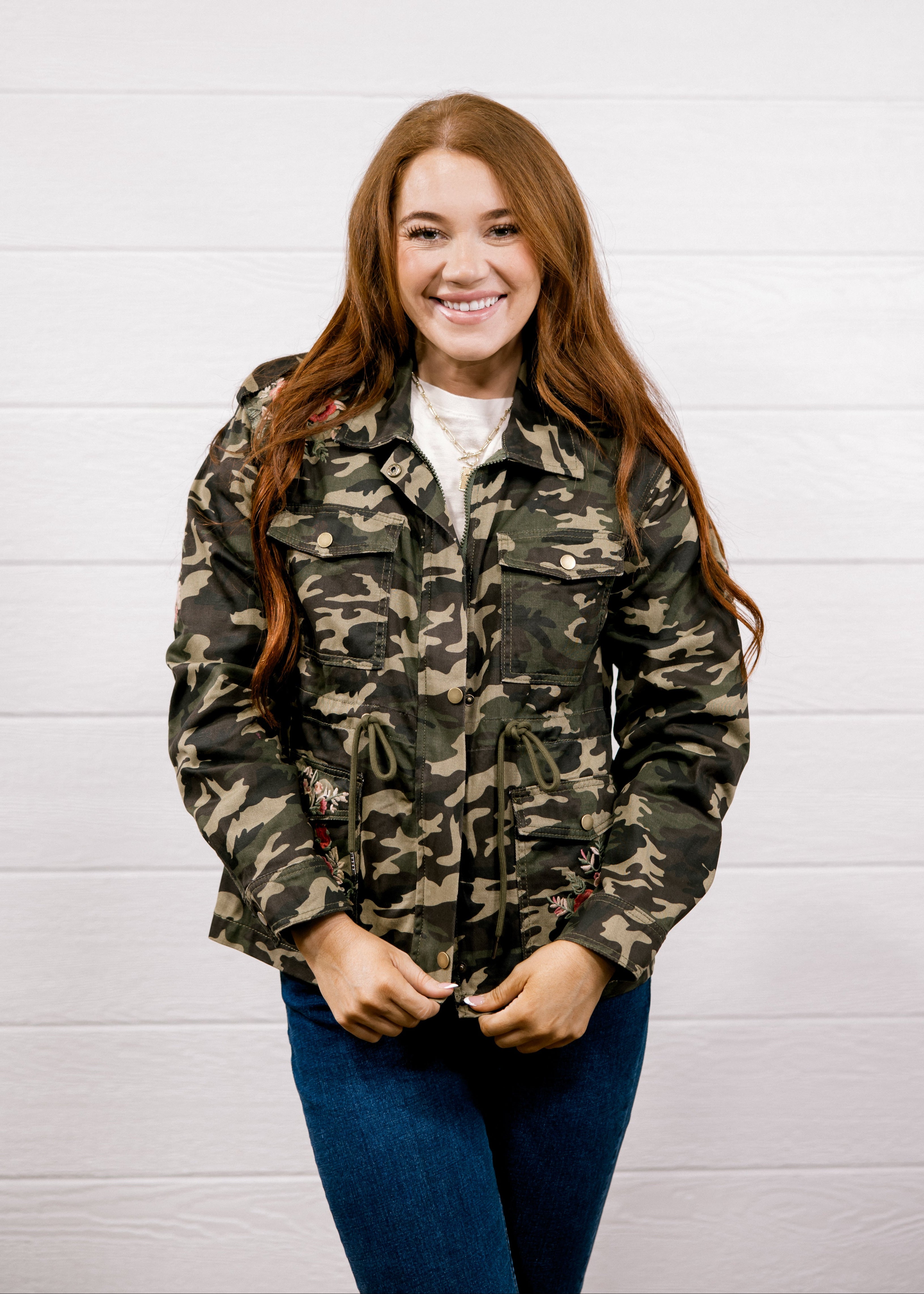 A smiling young woman with long red hair stands against a white paneled wall wearing a green and brown camouflage jacket over a white shirt and blue jeans.