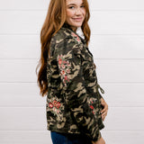 A woman with long light brown hair is shown from the side and back, wearing a camouflage jacket with red and pink floral embroidery, paired with dark blue jeans, against a white wood-paneled wall.