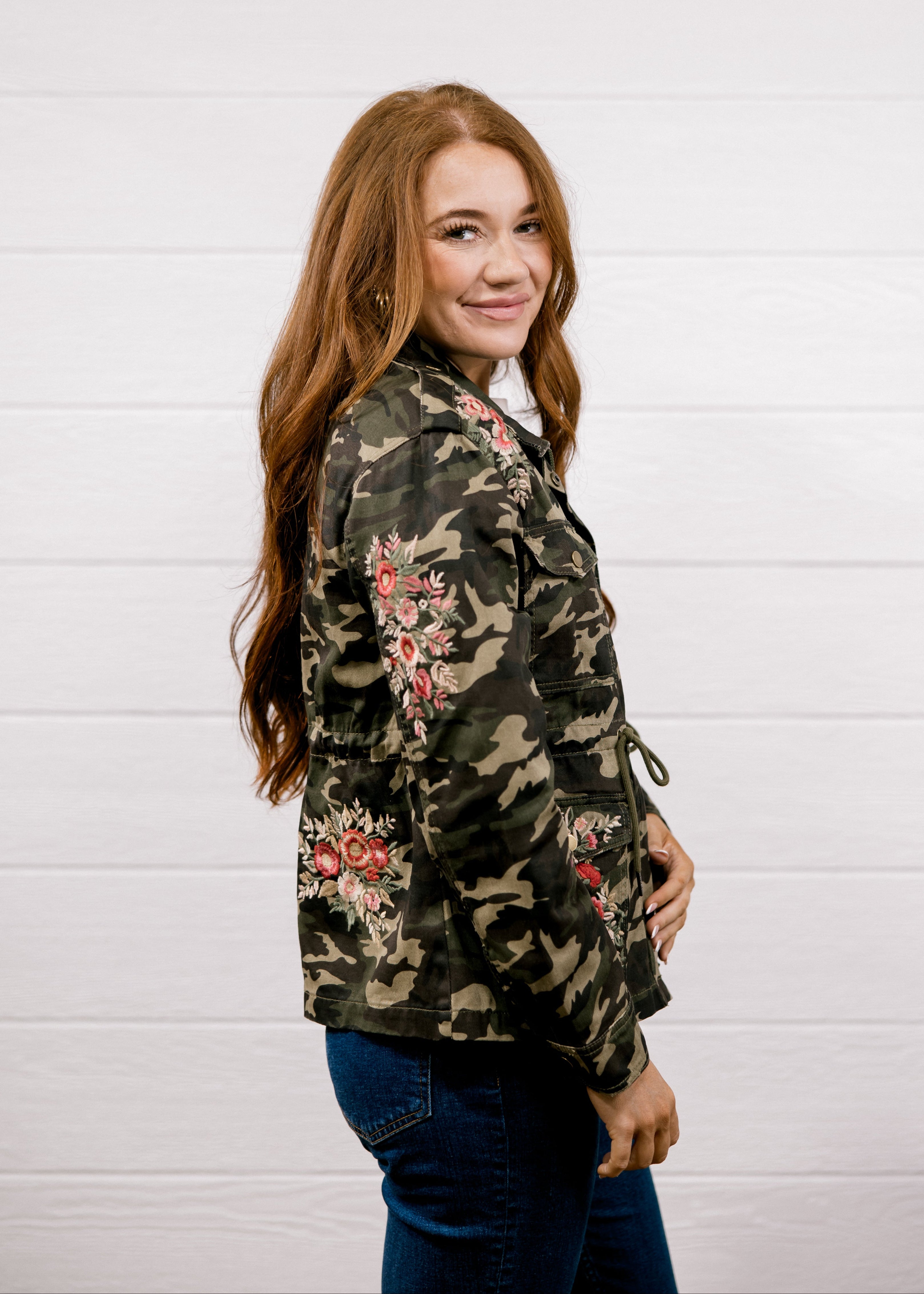 A woman with long light brown hair is shown from the side and back, wearing a camouflage jacket with red and pink floral embroidery, paired with dark blue jeans, against a white wood-paneled wall.