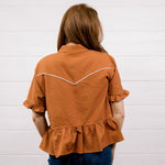 A woman viewed from behind wearing a rust-orange textured short-sleeve blouse with white contrast stitching across the upper back and dark blue jeans, standing against a white paneled wall.