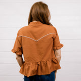 A woman viewed from behind wearing a rust-orange textured short-sleeve blouse with white contrast stitching across the upper back and dark blue jeans, standing against a white paneled wall.