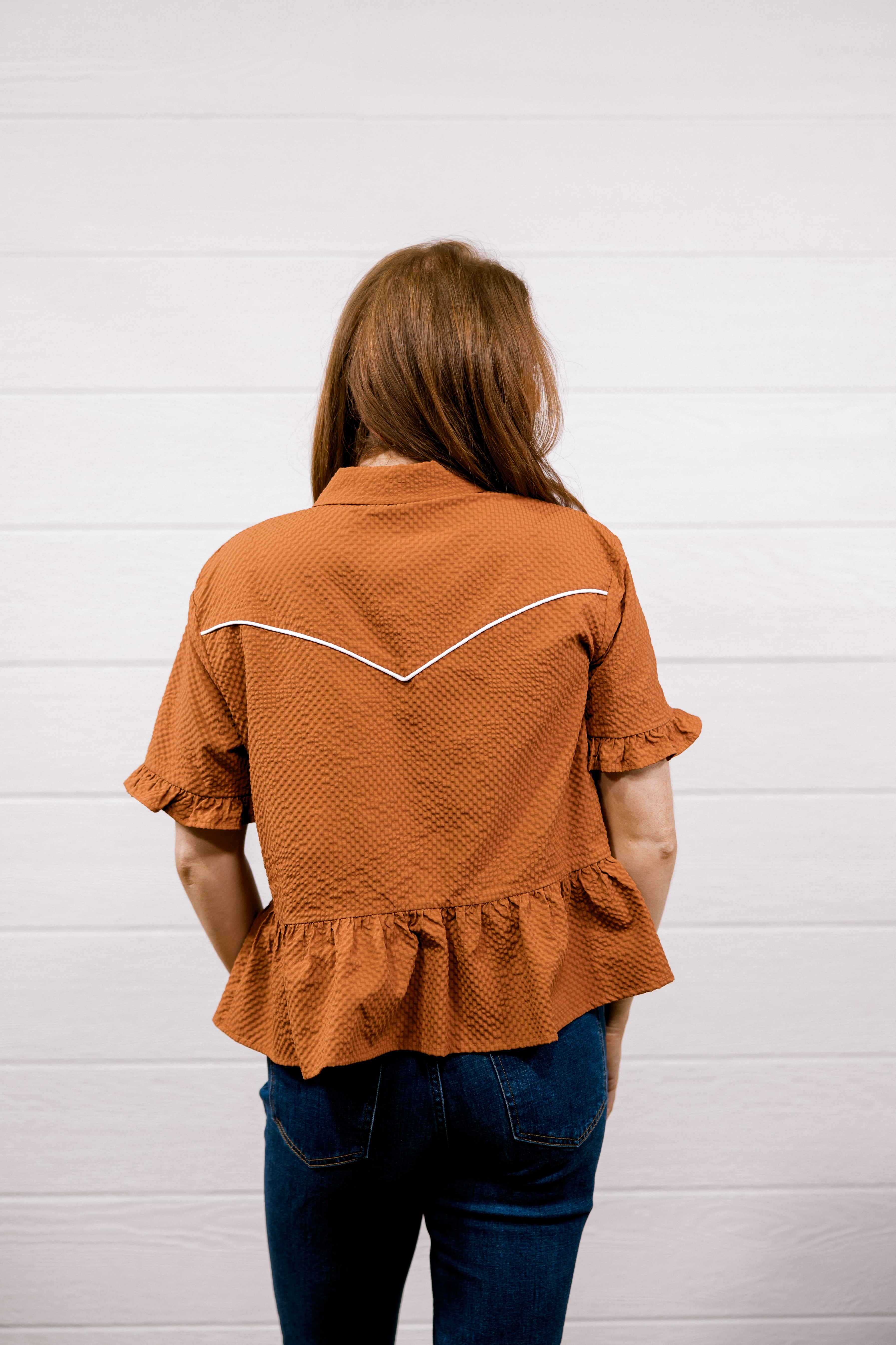 A woman viewed from behind wearing a rust-orange textured short-sleeve blouse with white contrast stitching across the upper back and dark blue jeans, standing against a white paneled wall.
