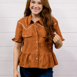 A smiling young woman with long brown hair stands against a white shiplap wall wearing a rust-orange short-sleeve western-style button-up shirt with white contrast stitching and dark blue jeans.