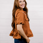 A woman with long brown hair wearing a rust-orange textured short-sleeve blouse and dark blue jeans, standing against a white wood-paneled wall.