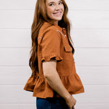 A woman with long brown hair wearing a rust-orange textured short-sleeve blouse and dark blue jeans, standing against a white wood-paneled wall.