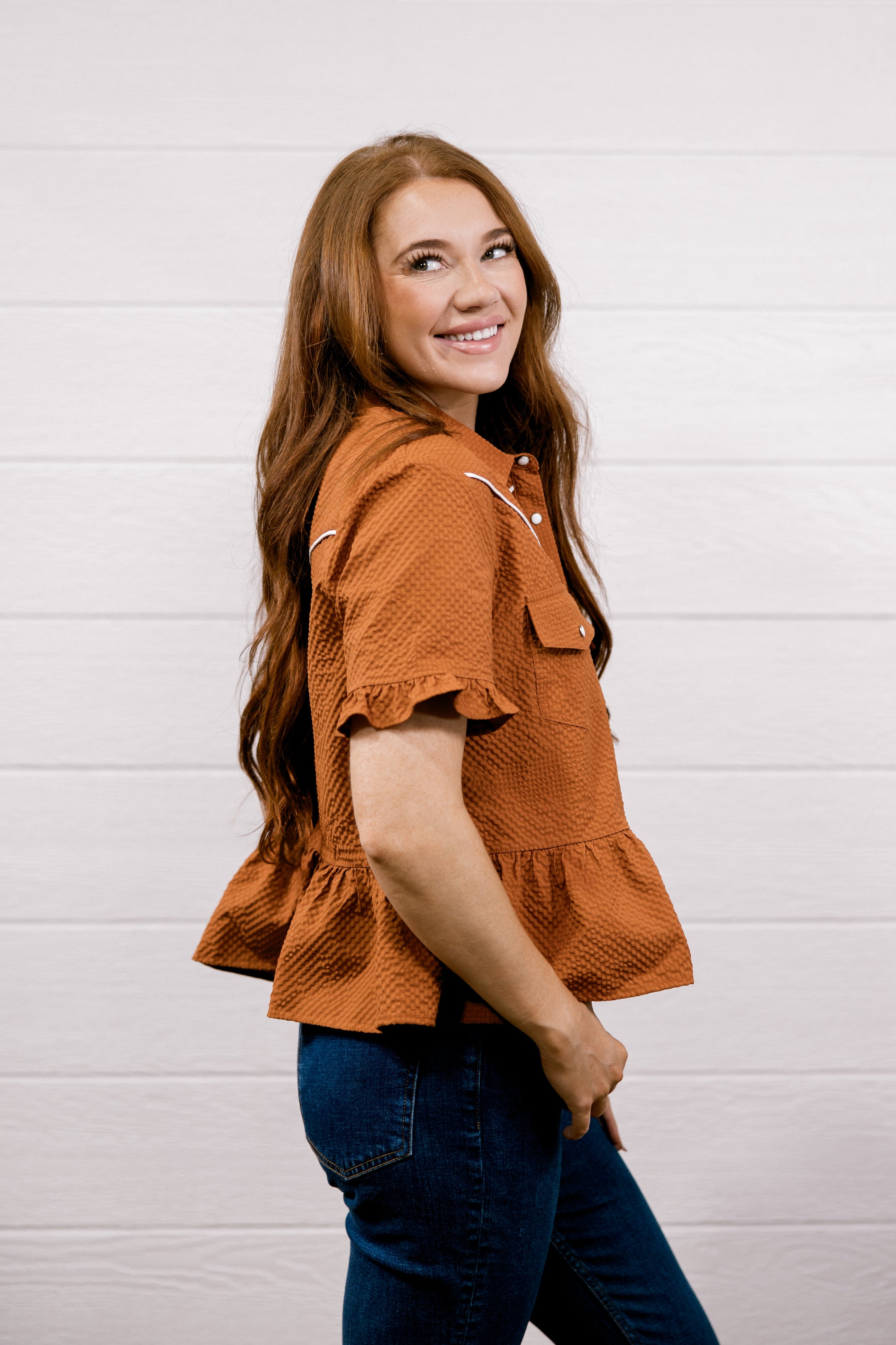 A woman with long brown hair wearing a rust-orange textured short-sleeve blouse and dark blue jeans, standing against a white wood-paneled wall.