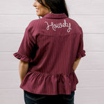 Woman wearing a maroon ruffled shirt with 'Howdy' embroidered on the back against a white background