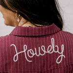 Close-up back view of a person wearing a burgundy pinstriped collared shirt or jacket with the word "Howdy" embroidered in white script across the upper back.