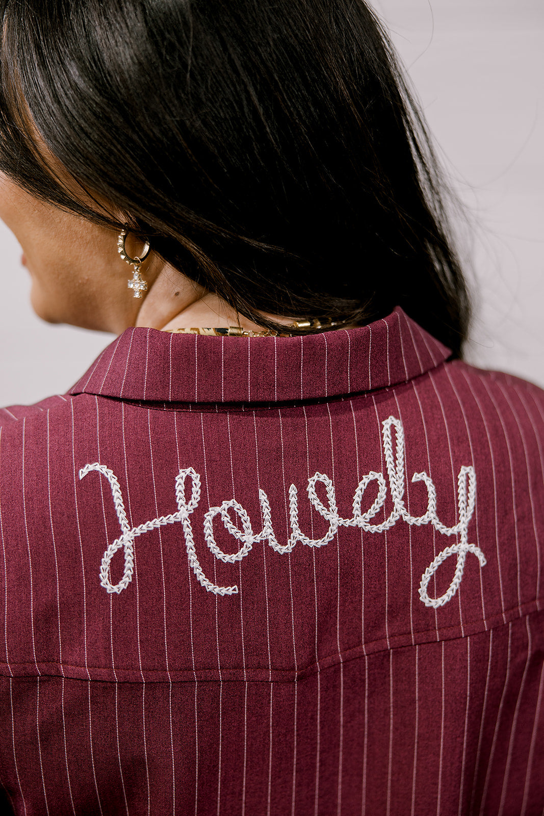 Close-up back view of a person wearing a burgundy pinstriped collared shirt or jacket with the word "Howdy" embroidered in white script across the upper back.