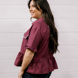 A woman with long dark hair is shown from the side/back wearing a short-sleeve ruffled blouse with thin vertical stripes in burgundy and white, paired with dark jeans, against a white paneled wall.