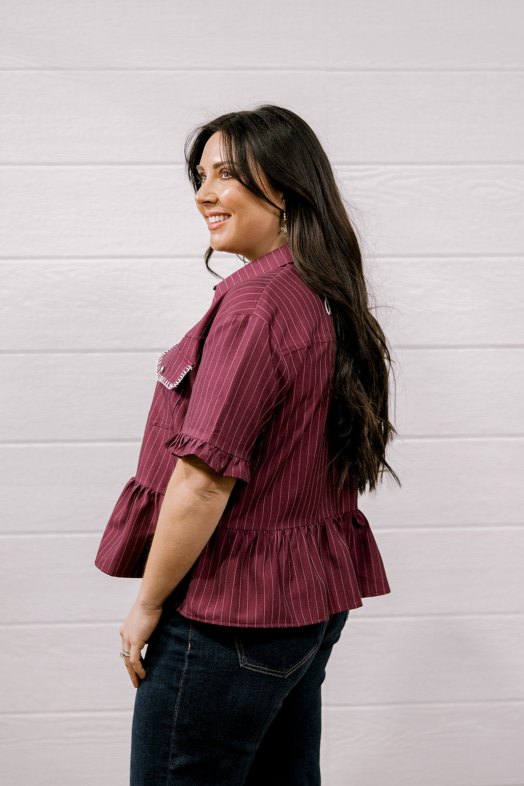 A woman with long dark hair is shown from the side/back wearing a short-sleeve ruffled blouse with thin vertical stripes in burgundy and white, paired with dark jeans, against a white paneled wall.