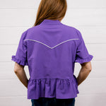 A woman viewed from behind wearing a purple short-sleeve blouse with white contrast stitching across the upper back and dark blue jeans, standing against a white paneled wall.