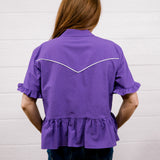 A woman viewed from behind wearing a purple short-sleeve blouse with white contrast stitching across the upper back and dark blue jeans, standing against a white paneled wall.