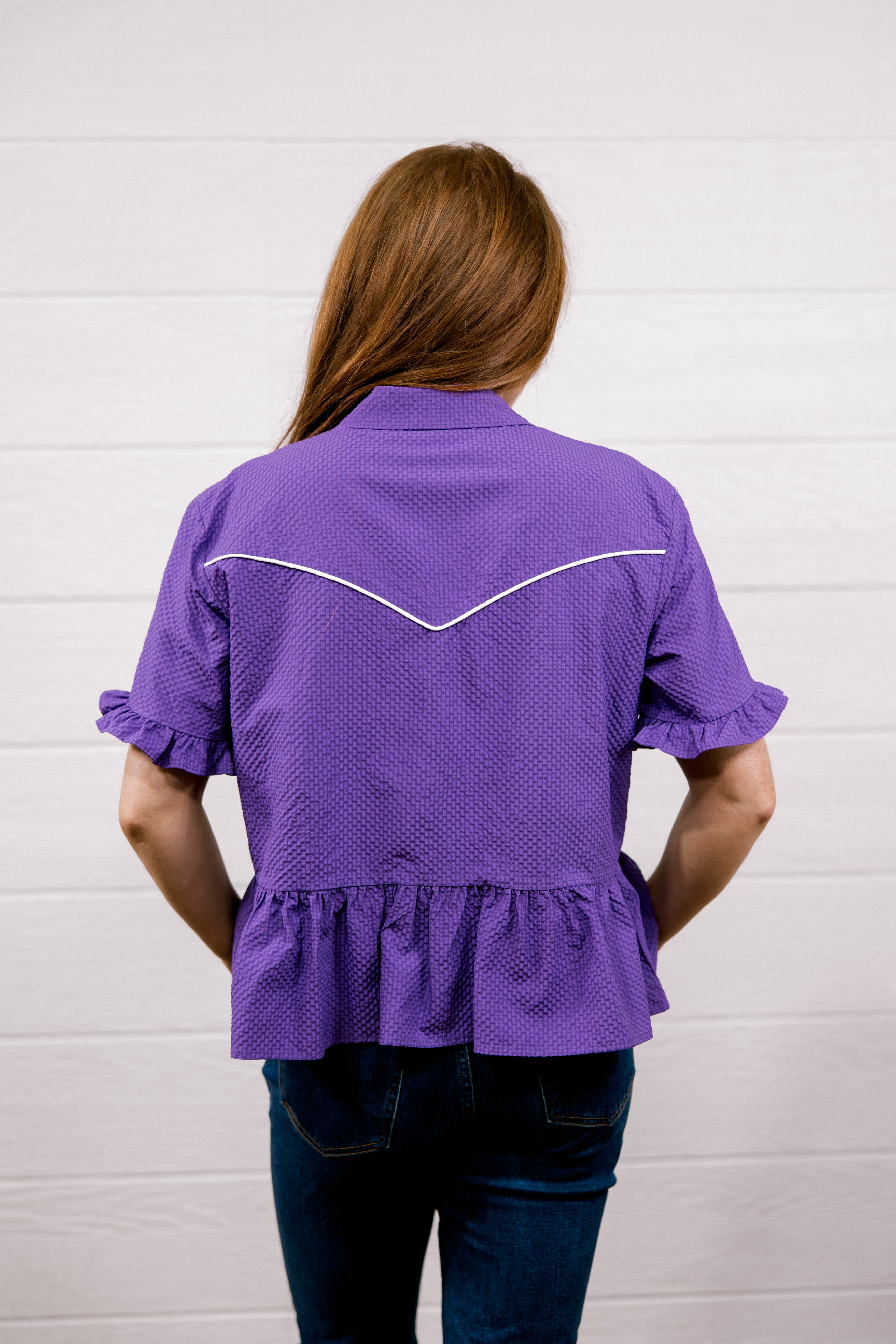 A woman viewed from behind wearing a purple short-sleeve blouse with white contrast stitching across the upper back and dark blue jeans, standing against a white paneled wall.