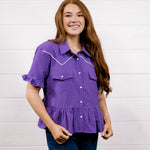 A woman with long light brown hair wearing a purple short-sleeve western-style blouse with white contrast stitching and dark blue jeans, standing against a white wood-paneled wall.