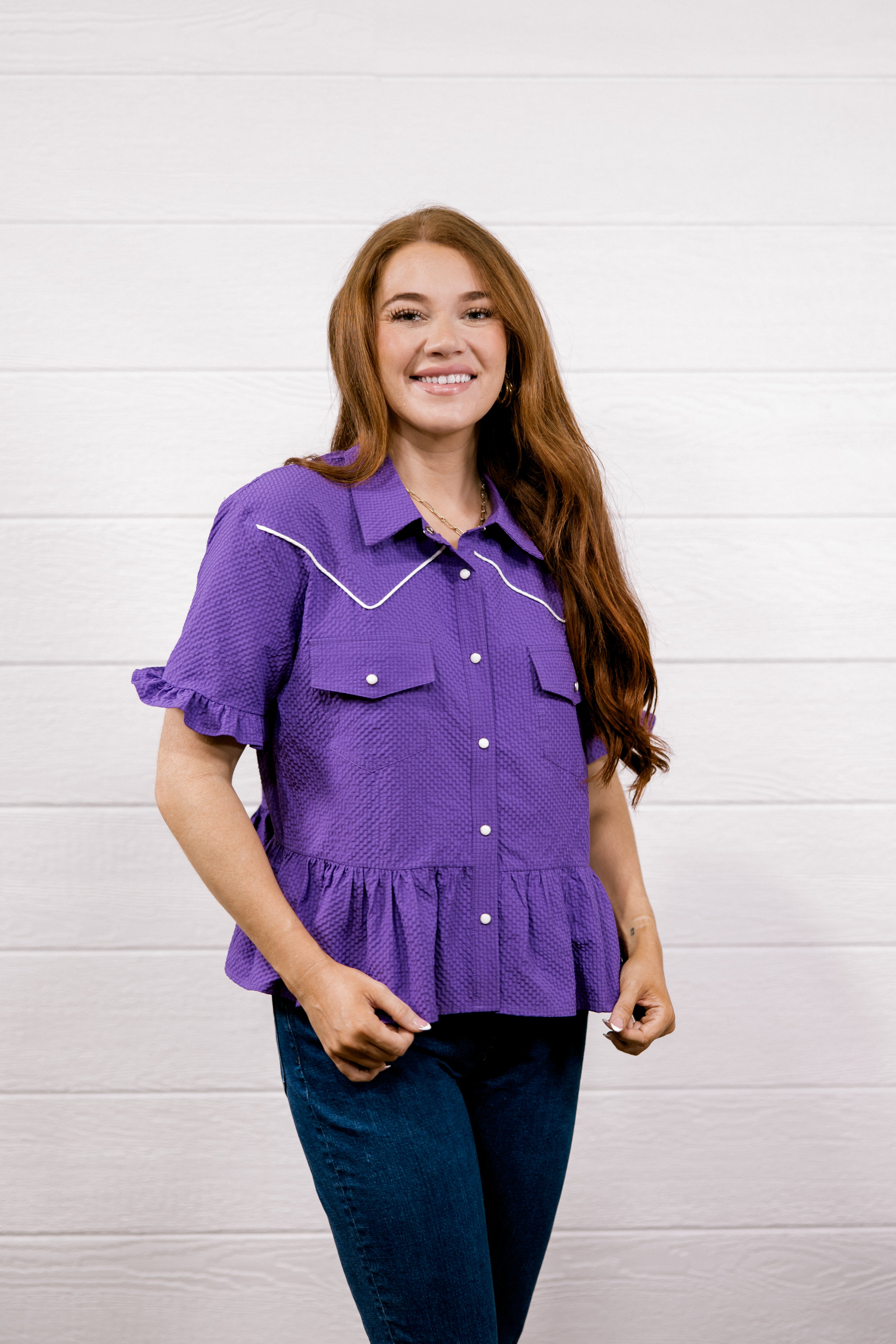A woman with long light brown hair wearing a purple short-sleeve western-style blouse with white contrast stitching and dark blue jeans, standing against a white wood-paneled wall.