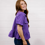 A woman with long light brown hair wearing a purple textured short-sleeve blouse with ruffle details and dark blue jeans, standing against a white wood-paneled wall.