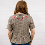 A woman viewed from behind wearing a brown and beige houndstooth short-sleeve blouse with floral embroidery across the upper back, paired with blue jeans, standing against a white wood-paneled wall.