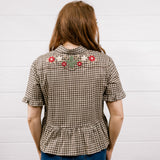 A woman viewed from behind wearing a brown and beige houndstooth short-sleeve blouse with floral embroidery across the upper back, paired with blue jeans, standing against a white wood-paneled wall.