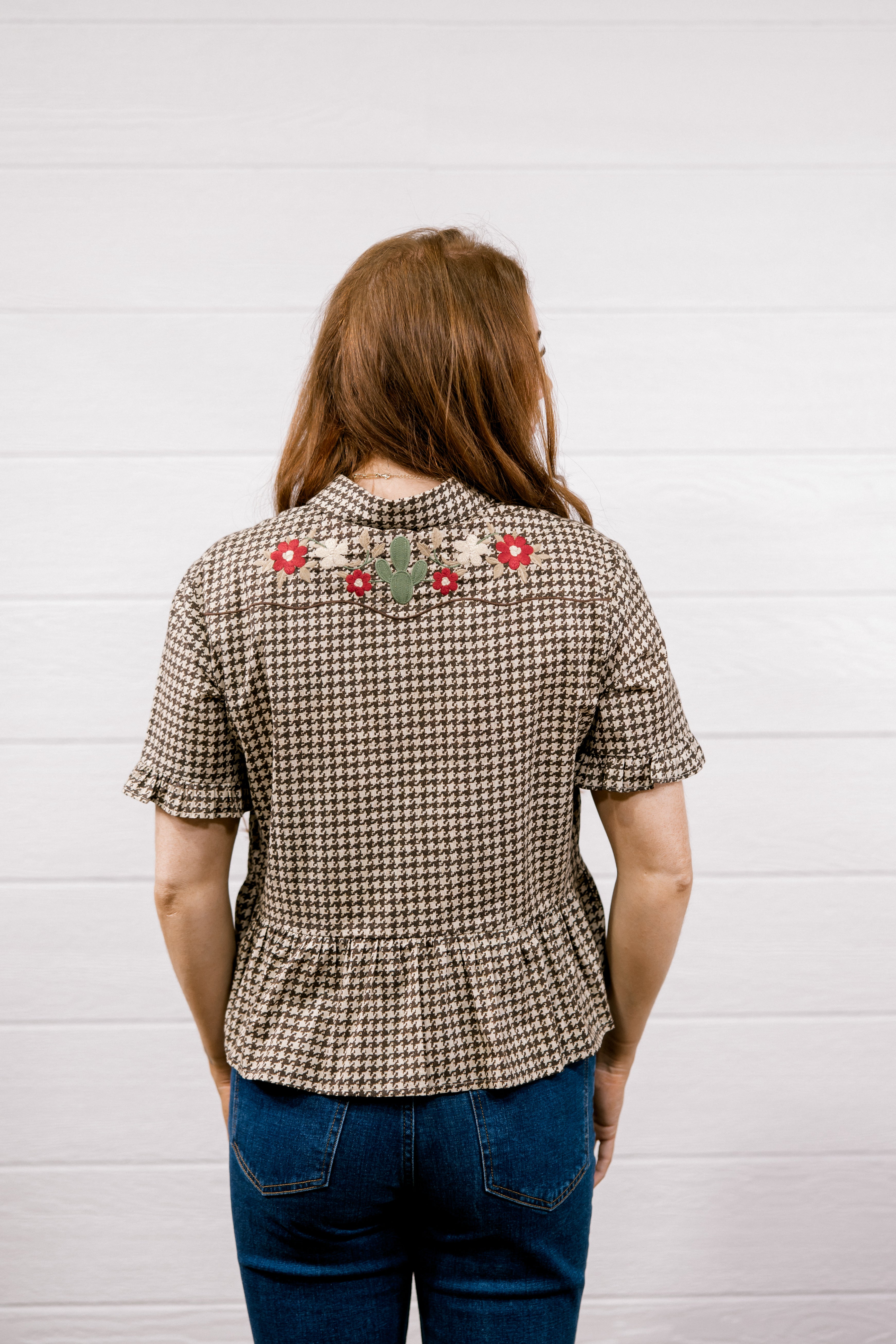 A woman viewed from behind wearing a brown and beige houndstooth short-sleeve blouse with floral embroidery across the upper back, paired with blue jeans, standing against a white wood-paneled wall.