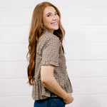 A woman with long wavy red hair is shown from the side and back, wearing a brown and beige houndstooth short-sleeve blouse and blue jeans, standing against a white wood-paneled wall.