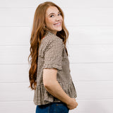 A woman with long wavy red hair is shown from the side and back, wearing a brown and beige houndstooth short-sleeve blouse and blue jeans, standing against a white wood-paneled wall.