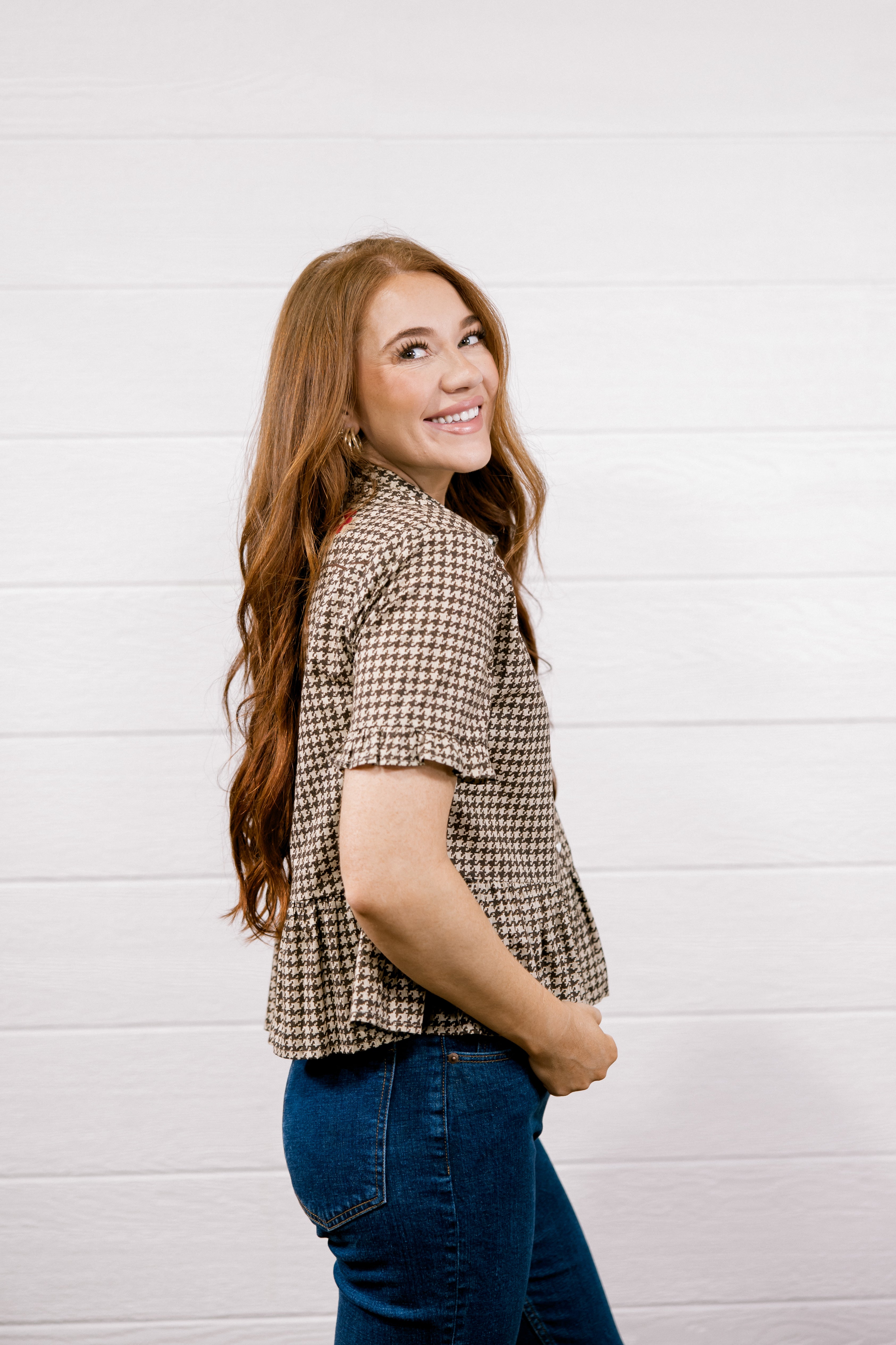 A woman with long wavy red hair is shown from the side and back, wearing a brown and beige houndstooth short-sleeve blouse and blue jeans, standing against a white wood-paneled wall.