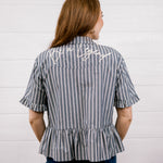 A woman viewed from behind wearing a short-sleeve striped blouse with vertical gray and white stripes, a peplum hem, and white script embroidery on the upper back. She is wearing dark blue jeans against a white wood-paneled wall.