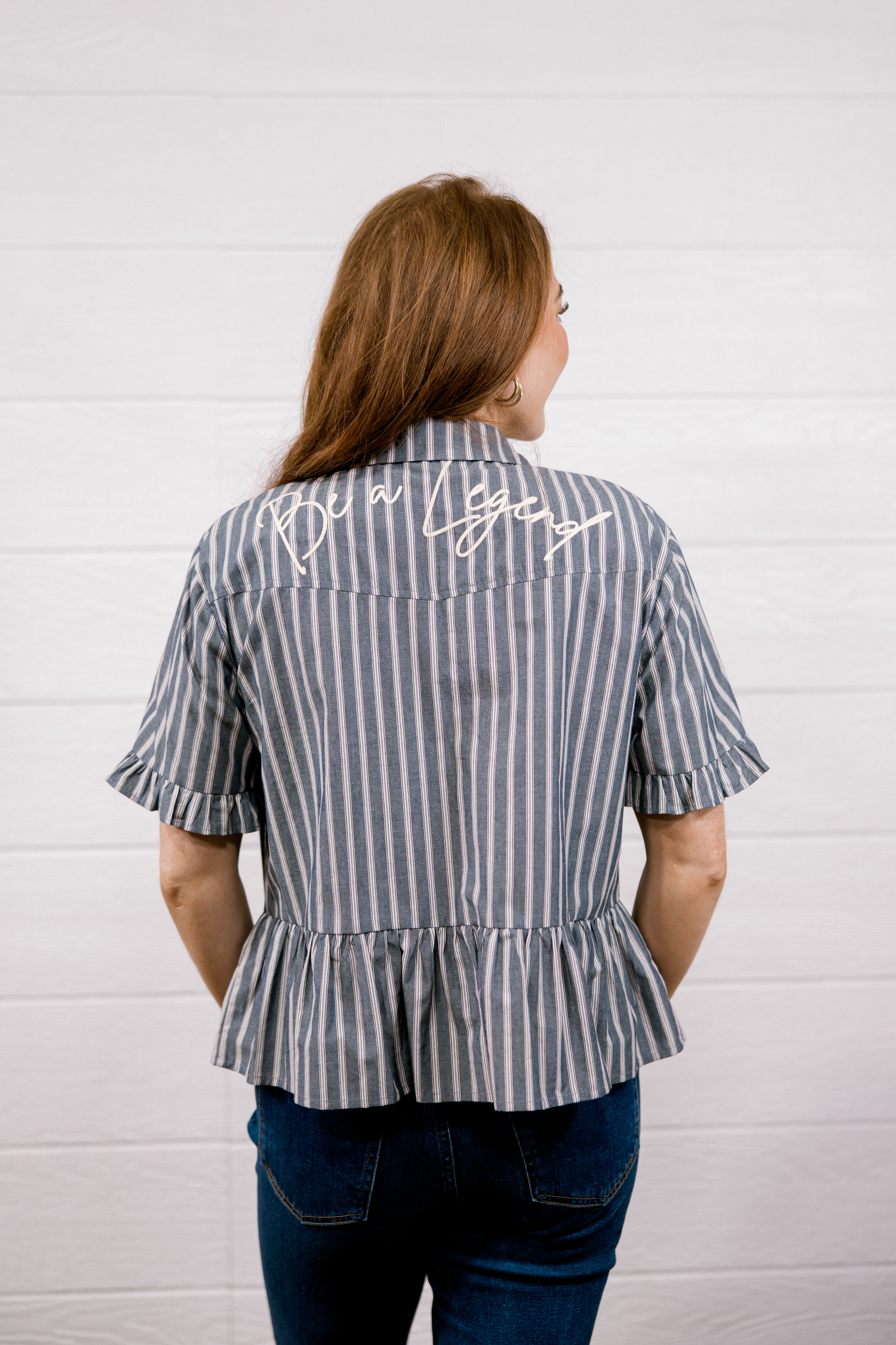 A woman viewed from behind wearing a short-sleeve striped blouse with vertical gray and white stripes, a peplum hem, and white script embroidery on the upper back. She is wearing dark blue jeans against a white wood-paneled wall.