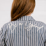 A woman viewed from behind wearing a blue and white vertically striped button-down shirt with 'Be a Legend' embroidered in white script across the upper back.