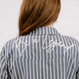A woman viewed from behind wearing a blue and white vertically striped button-down shirt with 'Be a Legend' embroidered in white script across the upper back.