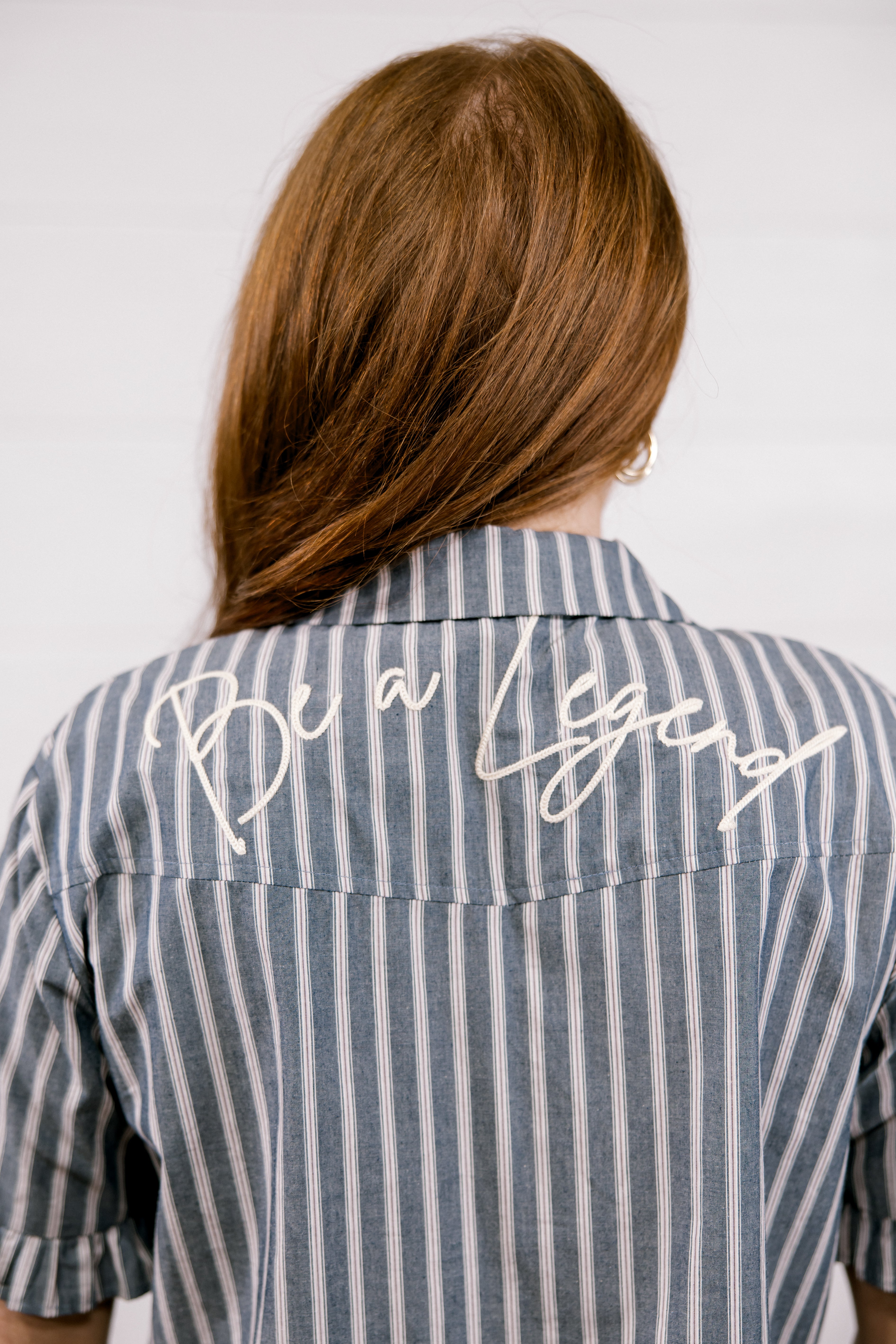 A woman viewed from behind wearing a blue and white vertically striped button-down shirt with 'Be a Legend' embroidered in white script across the upper back.