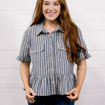 A smiling young woman with long red hair stands against a white shiplap wall wearing a short-sleeve button-front blouse with vertical gray and white stripes, ruffled sleeve cuffs, and two chest pockets, paired with dark blue jeans.