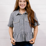 A smiling young woman with long red hair stands against a white shiplap wall wearing a short-sleeve button-front blouse with vertical gray and white stripes, ruffled sleeve cuffs, and two chest pockets, paired with dark blue jeans.