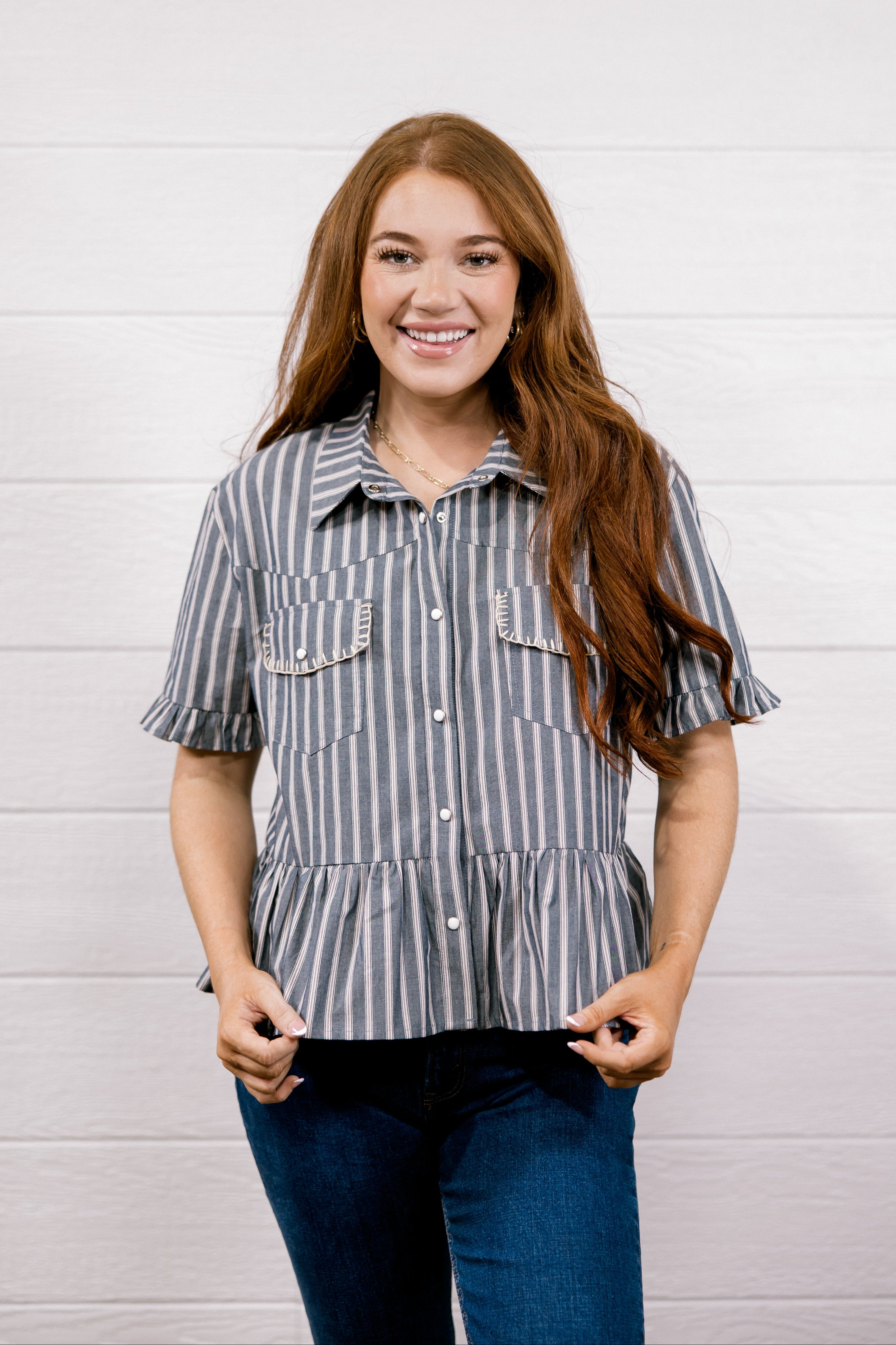 A smiling young woman with long red hair stands against a white shiplap wall wearing a short-sleeve button-front blouse with vertical gray and white stripes, ruffled sleeve cuffs, and two chest pockets, paired with dark blue jeans.