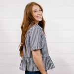 A woman with long light brown hair is shown from the back-three-quarter view, wearing a short-sleeve gray and white vertically striped ruffled blouse and dark blue jeans, standing against a white wood-paneled wall.