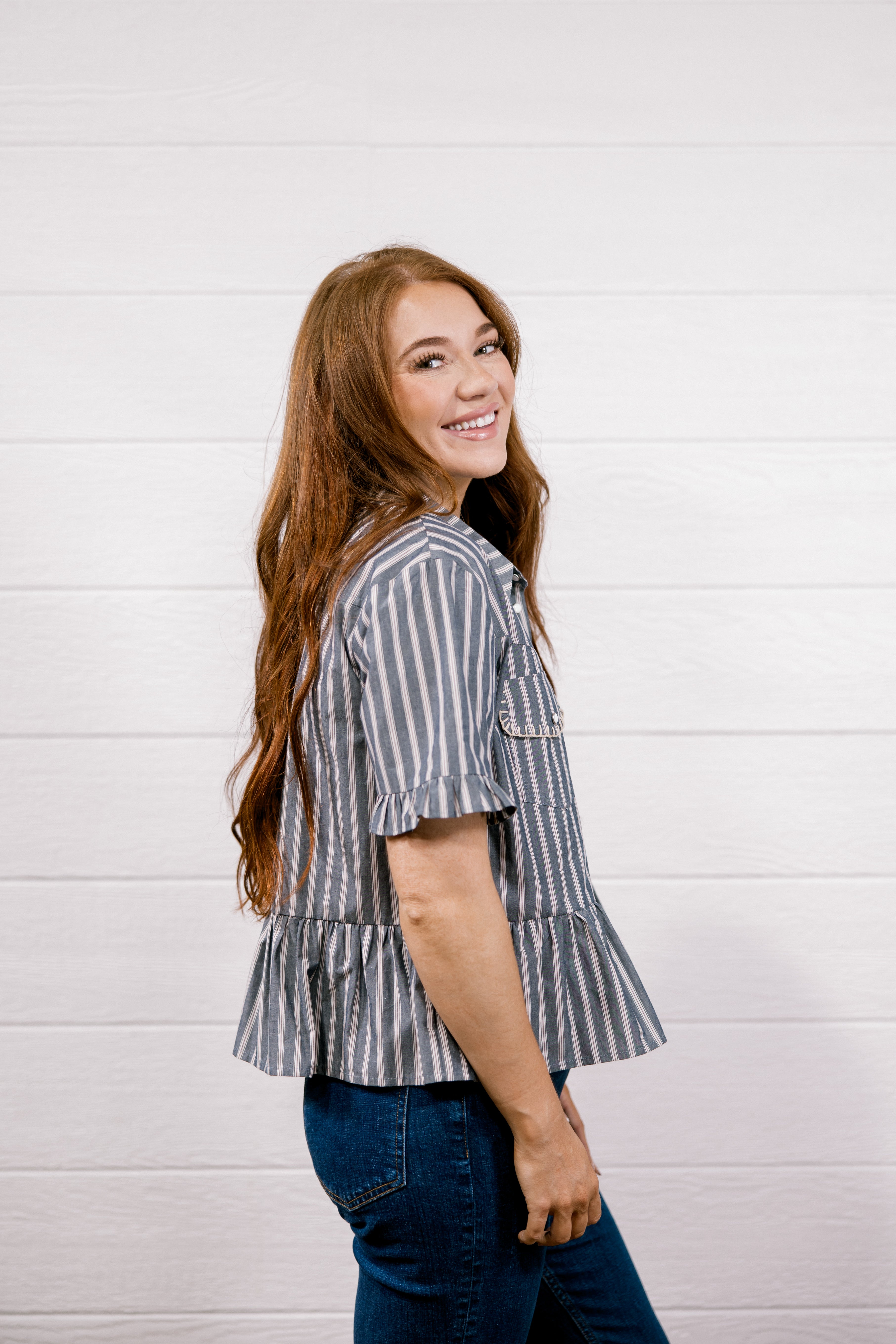 A woman with long light brown hair is shown from the back-three-quarter view, wearing a short-sleeve gray and white vertically striped ruffled blouse and dark blue jeans, standing against a white wood-paneled wall.