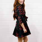 A woman modeling a black wrap-style mini dress with a red floral pattern, paired with white cowboy boots, standing against a white shiplap wall and wood floor.