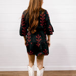 A woman viewed from behind wearing a black short dress with red floral embroidery and white cowboy boots, standing against a white paneled wall and wood floor.