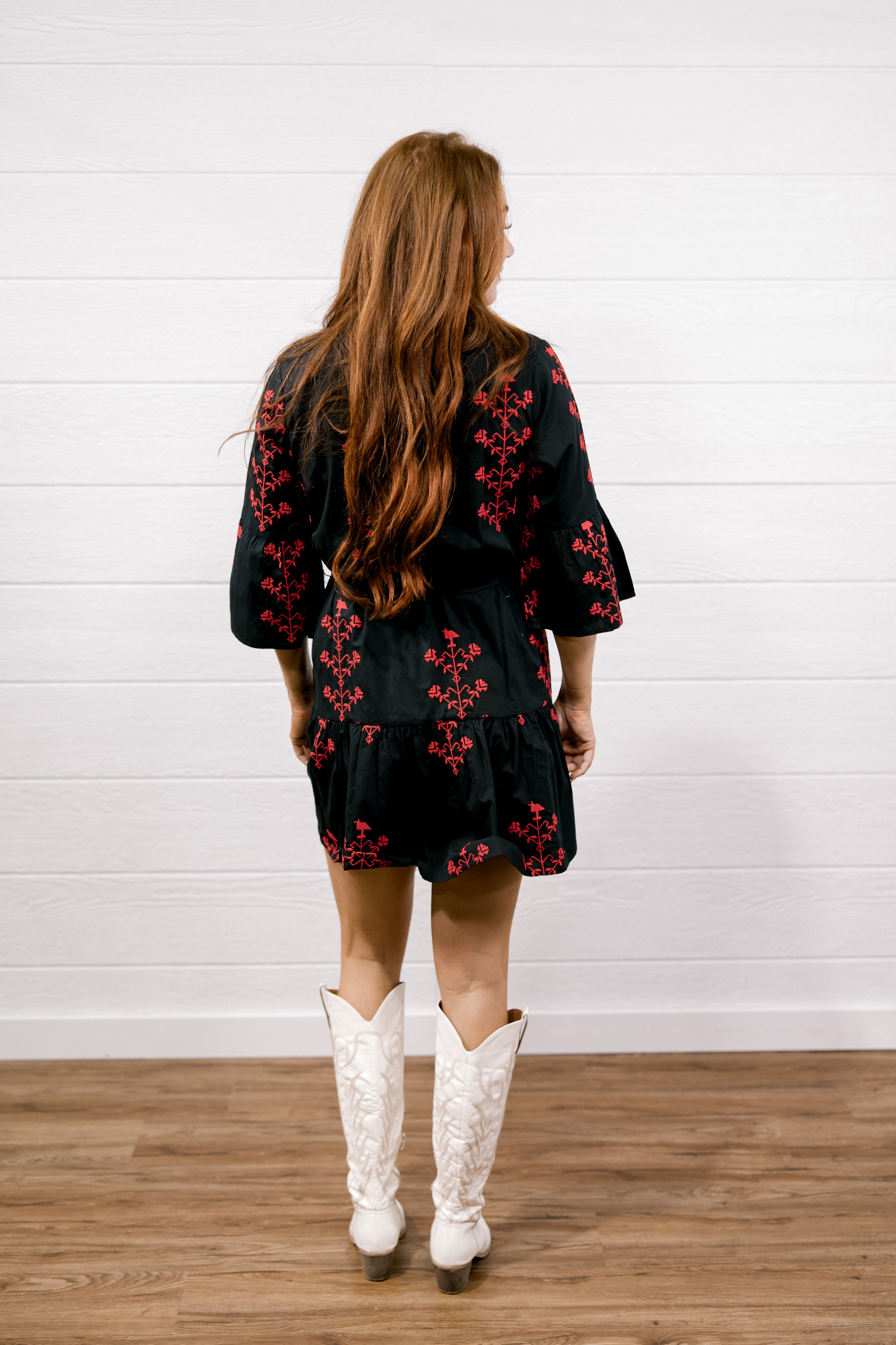 A woman viewed from behind wearing a black short dress with red floral embroidery and white cowboy boots, standing against a white paneled wall and wood floor.
