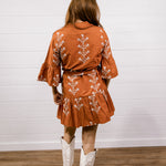 A woman viewed from behind wearing a rust-orange short dress with white floral embroidery, paired with white cowboy boots, standing against a white shiplap wall and wood floor.