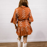 A woman viewed from behind wearing a rust-orange short dress with white floral embroidery, paired with white cowboy boots, standing against a white shiplap wall and wood floor.