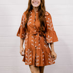 Woman wearing a rust-colored floral dress and white boots standing against a white wooden panel background.