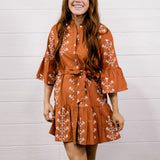 Woman wearing a rust-colored floral dress and white boots standing against a white wooden panel background.