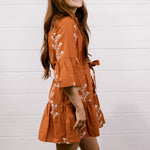 A woman modeling a rust-orange tiered mini dress with white floral embroidery and white cowboy boots, standing against a white shiplap wall on a wooden floor.