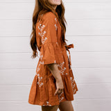 A woman modeling a rust-orange tiered mini dress with white floral embroidery and white cowboy boots, standing against a white shiplap wall on a wooden floor.