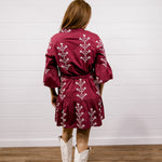 A woman viewed from behind wearing a burgundy mini dress with white floral embroidery, paired with white cowboy boots, standing against a white shiplap wall and wood floor.